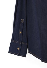 Mørk denimskjorte med en knappet ærmekant med synlige sømme og en buet kant. Detaljer inkluderer messingknapper og kontrasterende gul tråd.