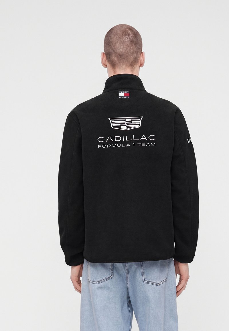 Човек с черно яке с лого на отбора Cadillac Formula 1 на гърба и светлосини дънки, стоящ с гръб към камерата.