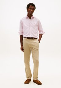 Man die met zijn hand in zijn zak staat, gekleed in een lichtroze gestreept overhemd met knopen, beige broek, bruine riem en bruine suède schoenen tegen een witte achtergrond.