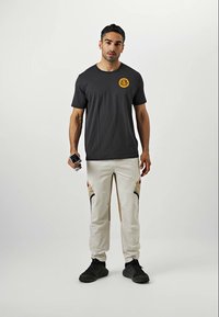 Homme se tenant contre un fond blanc, portant un t-shirt noir avec un logo circulaire jaune, un pantalon beige avec des accents noirs et des baskets noires, tenant un appareil photo.