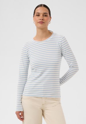 Frau mit dunklen Haaren im niedrigen Pferdeschwanz, die ein hellblau-weiß gestreiftes Langarmshirt und beige Hose trägt, steht mit einer Hand hinter dem Rücken.