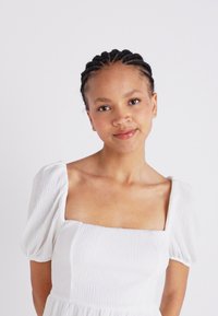 Λευκό, υφασμένο φόρεμα με τετράγωνη λαιμόκοψη και πλισέ μανίκια. Το ύφασμα φαίνεται ελαφρύ και αναπνεύσιμο, δημιουργώντας μια ανάλαφρη σιλουέτα.