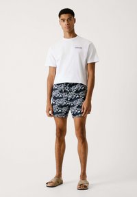 Camiseta blanca de algodón, pantalones cortos de baño azul marino con un patrón repetido de texto "Red Bull Racing" y sandalias beige tipo slider. Un atuendo simple y casual de verano.