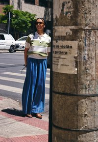 Kvinne med flettet hår og solbriller står på fortauet nær et fotgjengerfelt, iført en stripete skjorte og vide blå bukser.