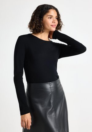 Kvinde med krøllet hår iført en sort langærmet top og sort lædernederdel, poserer med den ene hånd på sin hals og kigger mod højre.