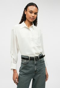 Valge pika varrukaga nööpidega särk, millel on rinna tasku, koos kõrge taljega siniste teksade ja musta vööga. Pehme, sile kangas.