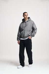 Young man standing wearing a gray zip-up hoodie, black jeans, white sneakers, looking right against a plain light background.