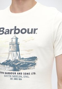Λευκό T-shirt με λογότυπο Barbour, γραφικό φάρου σε βράχια και κείμενο για την John Barbour and Sons Ltd., South Shields, Αγγλία, ιδρυθείσα το 1894.