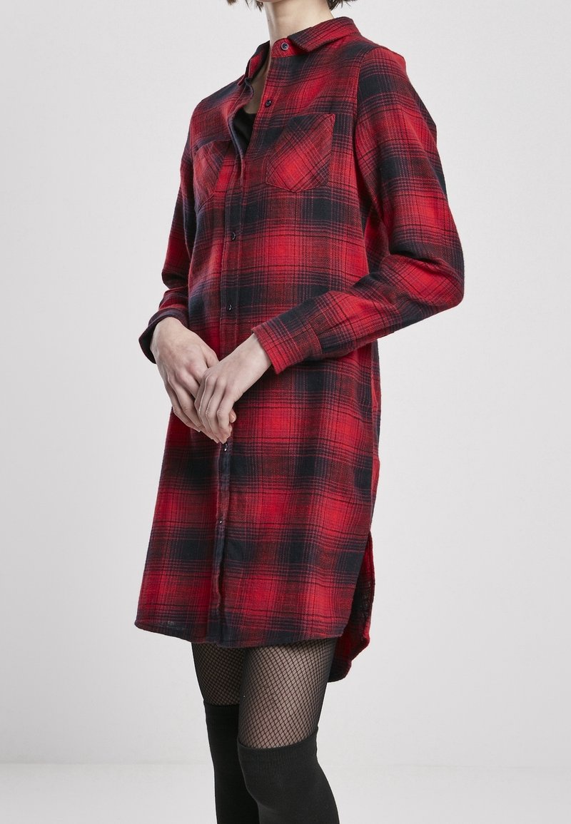 Femme portant une longue robe chemise à carreaux rouges et noirs avec des boutons à l'avant, associée à des collants résille noirs et des chaussettes noires au-dessus du genou.
