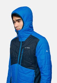 Homme portant une veste matelassée à capuche bleue et bleu marine avec le logo VAUDE, regardant légèrement vers le bas et sur le côté.