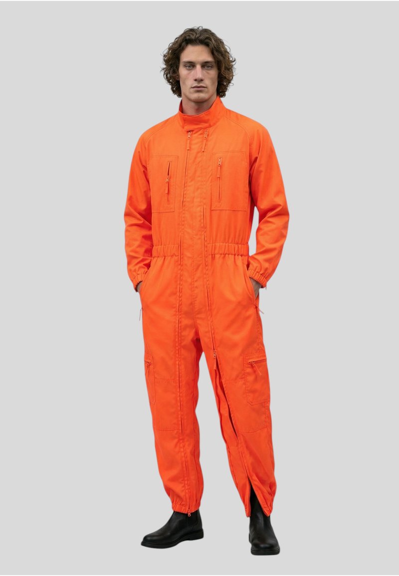 Homme aux cheveux bouclés portant une combinaison orange à fermeture éclair avec plusieurs poches et des bottines noires, debout les mains dans les poches.