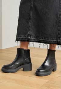 Botines de cuero negro hasta el tobillo con un tacón grueso, que cuentan con una cremallera lateral y una textura suave. Combinados con una falda negra con dobladillo deshilachado.