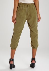 Persona vestida con pantalones joggers cortos de color verde oliva con puños elásticos y sandalias negras de tacón, de pie frente a un fondo liso.