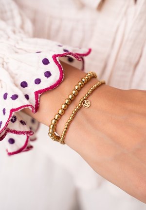 Bracciali d'oro su un polso, con due design con perline: uno liscio e rotondo, l'altro con un ciondolo. La manica ha un bordo arricciato con pois viola.