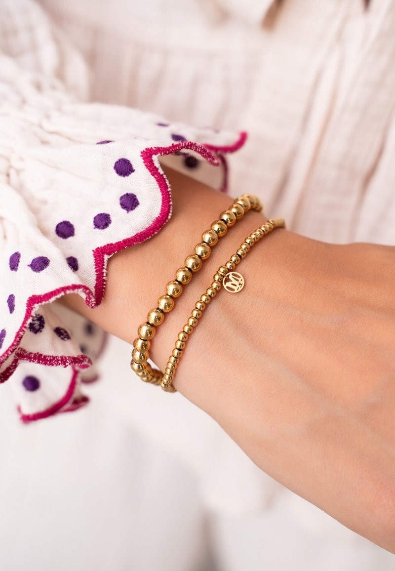 Bracciali d'oro su un polso, con due design con perline: uno liscio e rotondo, l'altro con un ciondolo. La manica ha un bordo arricciato con pois viola.