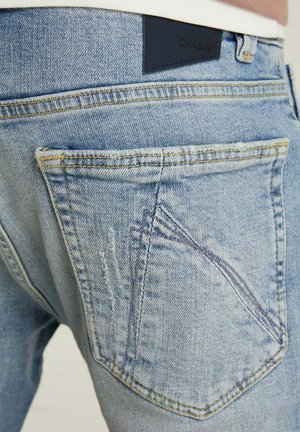 Lichtblauwe denim jeans met een verweerd textuur, voorzien van een donkere leren patch met de tekst "CHASIN'" op de achterste tailleband en schuin geplaatste achterzakken.