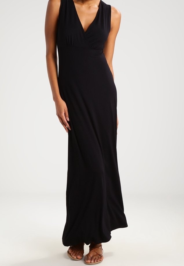Femme portant une robe noire sans manches longue jusqu'au sol avec un col en V, associée à des sandales plates marron à brides.