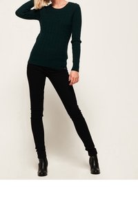 Dunkelgrüner gerippter Strickpullover mit langen Ärmeln, kombiniert mit schwarzen engen Hosen und schwarzen Ankle Boots, stehend vor einem hellen Hintergrund.