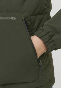 Grøn puffjakke med en lynlås lomme, elastiske mansjetter og en mat finish. Har logoen "Jack & Jones" trykt tæt på lommen.