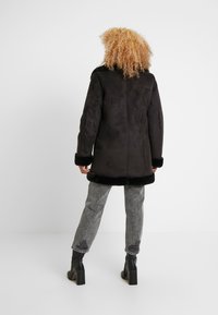 Zwarte jas met kunstbontvoering, lange mouwen en een gestructureerde stof. Heeft een zwarte bontafwerking bij de manchetten en zoom. G gedragen met versleten grijze jeans.