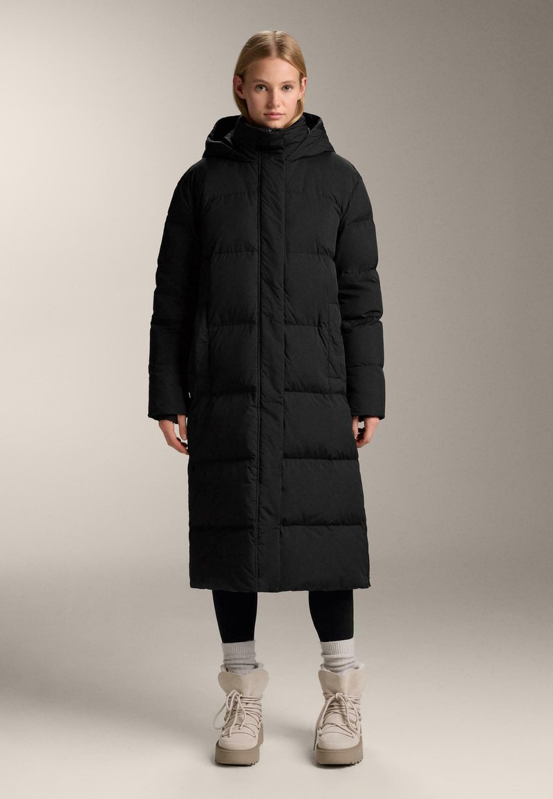 Fekete steppelt kabát kapucnival, hosszú ujjakkal és egyenes szabással. Fekete leggings és vastag bézs csizmák fölött viselve.