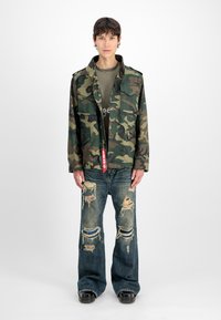 Joven vistiendo una chaqueta de camuflaje, camiseta verde, jeans anchos rasgados y zapatos negros gruesos, de pie contra un fondo blanco liso.