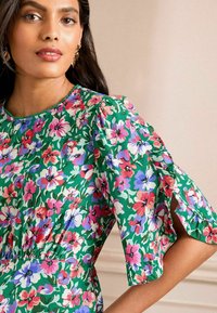 Ziedu kleita ar zaļu fonu, uz kura redzami rozā, purpura un baltie ziedi. Īsām piedurknēm ir šķelšanās detaļas. Gluda auduma tekstūra.