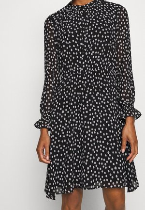 Robe noire à manches longues avec des pois blancs irréguliers, fermeture à boutons sur le devant, taille élastique et poignets à volants, portée par une personne.