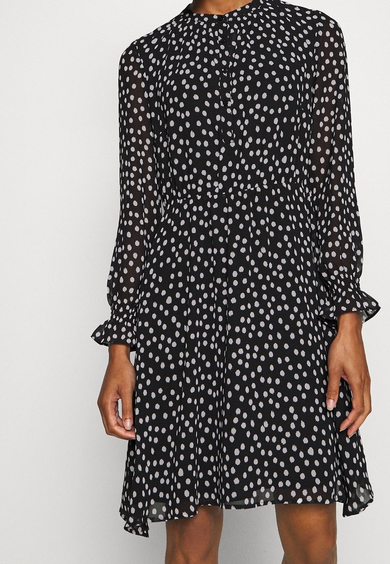 Robe noire à manches longues avec des pois blancs irréguliers, fermeture à boutons sur le devant, taille élastique et poignets à volants, portée par une personne.