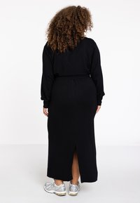 Schwarzes Maxikleid mit langen Ärmeln, einem elastischen Taillenbund und einem Schlitz im Rücken, kombiniert mit silbernen Sportschuhen. Weicher Stoff.