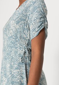 Blå och vit mönstrad klänning med korta rynkade ärmar, v-ringning och sidoknyttedetalj. Tyget har en slät, mjuk textur.