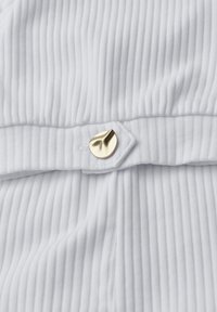 Tissu blanc côtelé avec un pli maintenu par un petit bouton doré brillant, de forme irrégulière, qui fixe le matériau.