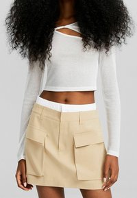 Haut court blanc à manches longues côtelé avec un col torsadé, associé à une mini jupe beige avec deux poches cargo avant.