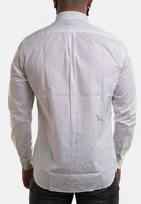 Camicia bianca a maniche lunghe con colletto standard, texture liscia e dettaglio con piega sul retro; abbinata a jeans scuri.