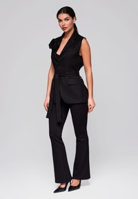 Zwarte mouwloze broekpak met een tailleband, voorzien van een revers en een bloemenaccent op de schouder, gecombineerd met uitlopende pantalons en puntige schoenen.