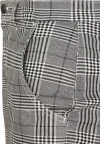 Gros plan sur un tissu à carreaux noir et blanc montrant la poche d'un pantalon avec des coutures diagonales et un bouton sur la ceinture.