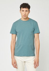 T-shirt a maniche corte color blu verde in un tessuto morbido, con collo a giro e taschino sul lato sinistro del petto. Abbinato a pantaloni chiari.