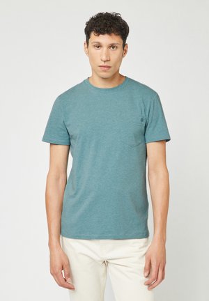 Teal korte-mouwen t-shirt van zachte stof, met een ronde hals en een borstzakje aan de linkerkant. Gestyled met lichtgekleurde broek.