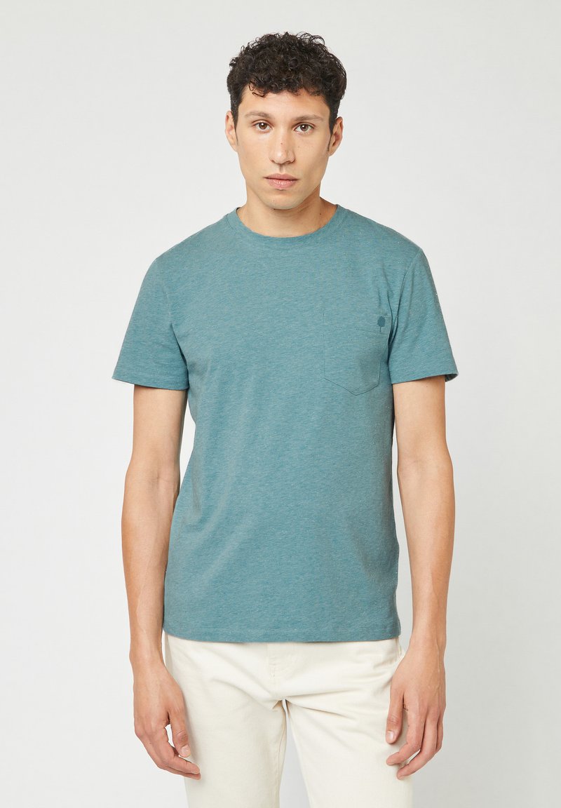 T-shirt a maniche corte color blu verde in un tessuto morbido, con collo a giro e taschino sul lato sinistro del petto. Abbinato a pantaloni chiari.