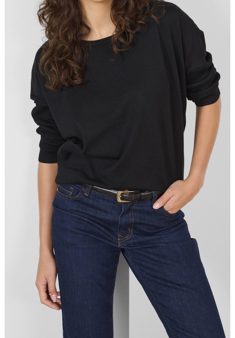 Pull noir en maille avec large encolure ronde et manches côtelées, associé à un jean bleu foncé avec des coutures bleu clair et une ceinture noire.