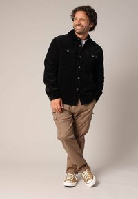 Zwarte corduroy jas met twee borstzakken, khaki cargobroek en beige sneakers met witte zolen. Model staat met een lichte buiging.
