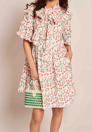 Woman wearing a white dress with red cherry pattern and ruffled sleeves, holding a small green and white beaded handbag.