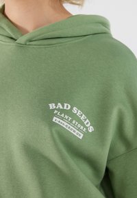 Grön hoodie i mjukt tyg, med vit text som läser "BAD SEEDS PLANT STORE" och en böjd grafisk design.