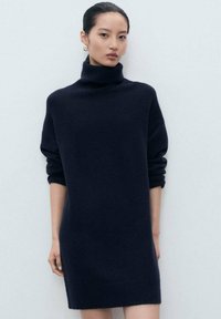 Donna che indossa un abito maglione a collo alto oversize blu navy con maniche lunghe arrotolate, in piedi contro uno sfondo chiaro e uniforme.