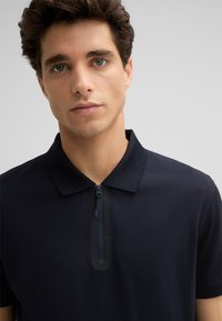 Młody mężczyzna z krótkimi brązowymi włosami, ubrany w granatową koszulkę polo z pół-zamkiem, zwrócony w stronę aparatu na jasnym, jednolitym tle.