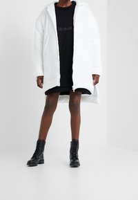 Biała pikowana kurtka z kapturem, zapinana na zamek z przodu. Pod spodem czarna sukienka koszulowa oraz czarne sznurowane botki z teksturowaną podeszwą.