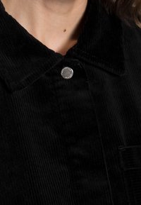 Camicia di corduroy nera con colletto, dotata di un bottone a pressione argento e texture a coste verticali. Nessuna tasca visibile nella vista accorciata.