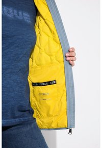 Chaqueta acolchada gris con un interior de un brillante color amarillo, que cuenta con un bolsillo interior etiquetado y una tela texturizada. Mano visible sosteniendo la chaqueta.
