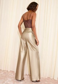 Donna che indossa pantaloni metallici larghi d'oro e una maglietta senza maniche marrone, in piedi con le spalle rivolte verso uno sfondo di tende chiare.