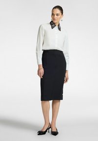 Witte blouse met een zwarte geborduurde kraag, gecombineerd met een aansluitende zwarte kokerrok met gouden knoopaccenten en zwarte slingbacks.